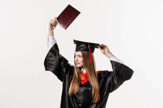 Happy girl student in black graduation gown and cap raises masters degree diploma above head on white background. Graduate girl is graduating college and celebrating academic achievement.