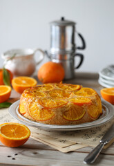 Torta d'arancia decorata con fette caramellate in primo piano. Sullo sfondo arance e utensili per colazione e caffè