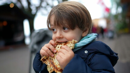 Small boy eating pancake outside in city street during winter season. Hungry child eating food during december festivities