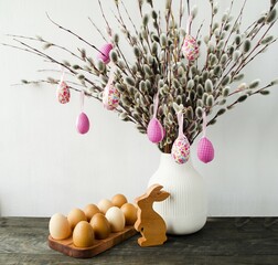 Decorative Easter composition in natural and ecological style.  On a wooden table stands a white vase with willow branches and Easter eggs, a wooden rabbit.  Front view, bright Easter holiday concept.