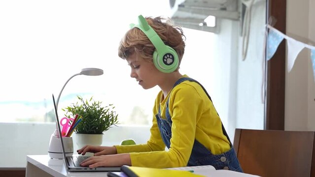 Cute smart caucasian primary School Child with headphones typing on the keyboard laptop. Online tutor teaching digital class, web lesson on computer at home. Virtual education. Schoolboy girl studying