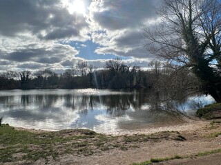 Landscape of Norfolk Broad with sandy shore by large lake , the Spring blue sky with sun light through white puffy cloud and trees reflected  in calm river water on bright cold day in East Anglia UK