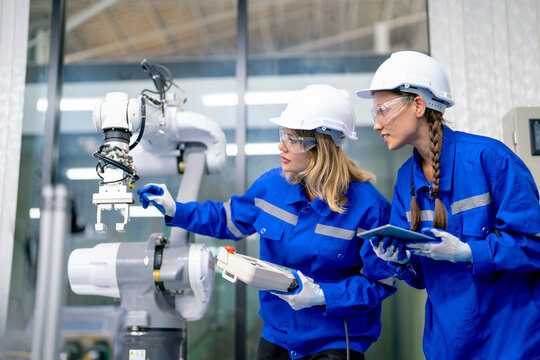 Two Caucasian professional engineer or technician woman workers hold controller and tablet help to check and maintenance robotic arm machine with stand beside in factory workplace.