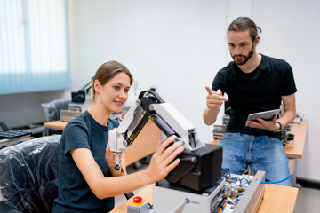 Caucasian professional engineer or technician manager man with casual cloth help to support and also guide staff to check small robotic machine that put on table in factory workplace.