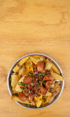 French fries. Top view of fried potatoes with sliced green onion, smoked sausages, crispy bacon and cheddar cheese sauce, in a metal dish on the wooden table.