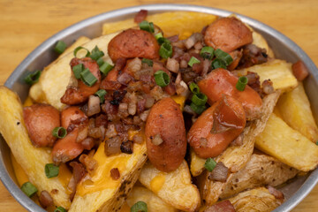 French fries. Top view of fried potatoes with sliced green onion, smoked sausages, crispy bacon and cheddar cheese sauce, in a metal dish on the wooden table.