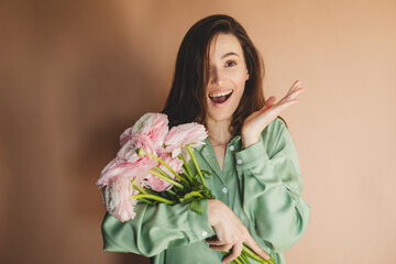 International Women's Day. Extremely happy woman in green shirt enjoying a bunch of spring flowers ranunculus, which she is holding in her hands. Wow emotions, hand near head. Surprised girl.
