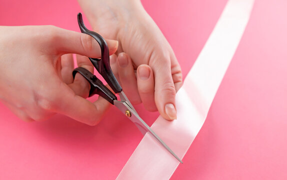 Scissors in female hands on a pink background cut a pink ribbon.