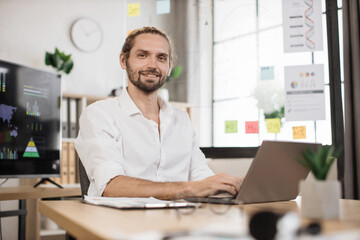 Fototapeta na wymiar Attractive positive caucasian guy in white shirt posing at own modern office. Young bearded businessman with shining smile, sitting at desk with papers and modern laptop.