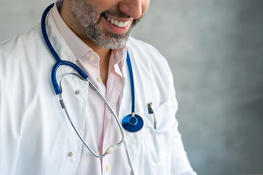 Close up crop photo of unrecognizable handsome smiling male doctor, hospital worker with beard or medical student in white lab medical coat having stethoscope around his neck and pen in coat pocket.