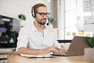 Busy caucasian man in white shirt and headset sitting at office and using wireless laptop. Big monitor with various charts and graphs on background. Business and finance concept.