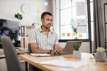 Busy african american man in striped shirt sitting at office and using wireless laptop. Big monitor with various charts and graphs on background. Business and finance concept.