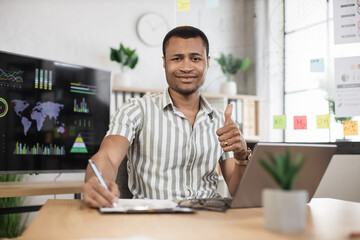 Fototapeta na wymiar Portrait of confident african businessman in stylish formal wear writing notes using laptop while sitting at modern office desk. Handsome company worker looking at camera showing thumbs up.
