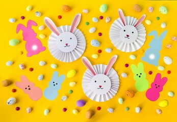 Craft, on a yellow background, cute, cute white paper rabbits and multi-colored Easter eggs.  Flat lay, top view.  Pattern, background picture, concept holiday light Easter.