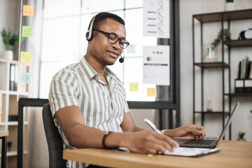 Fototapeta premium Portrait of smiling african young male office manager in striped shirt and headset writing financial report using wireless laptop computer during working online.