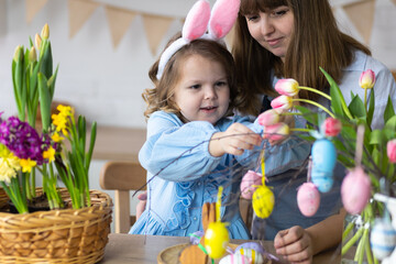 Mother and daughter painting eggs, creating home Easter decor together, happy, cheerful. Cute pretty smiling little girl, family time. Fresh spring bright colourful flowers in the kitchen. Sunny day