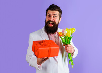 Smiling bearded man with gift box and bouquet of flowers. Businessman in suit with gift and bouquet of tulips. Romantic man with bouquet of flowers and present box. Valentines or Womens Day, birthday.