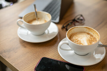 Two cups of fragrant cappuccino coffee stand on wooden table next to laptop. Coffee break. Online work in cafe. 