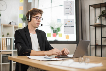 Young businessman in headset using laptop computer during video call working in office. Concentrated adult successful man wearing official suit sitting at wooden desk indoor
