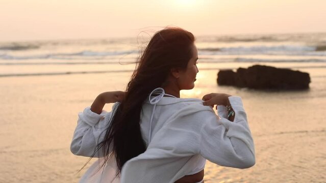 Happy Asian woman with long wavy hair in white shirt enjoys light breeze on ocean shore, exposing waters to wind. 
