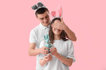 Young man with Easter rabbit closing her girlfriend's eyes on pink background
