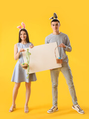 Happy young couple in bunny ears with Easter eggs and blank poster on yellow background
