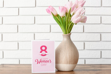 Vase with tulips and greeting card for Women's Day on table near white brick wall