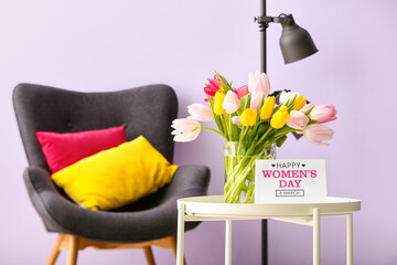 Vase with tulips, greeting card for Women's Day on table and armchair near lilac wall in room