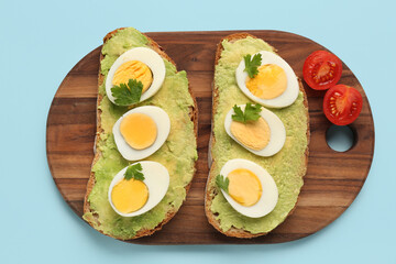 Board with delicious avocado toasts, boiled eggs, parsley and tomatoes on blue background