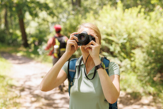 Diverse couple with backpacks walking in sunny forest, woman stopping to take photo with camera