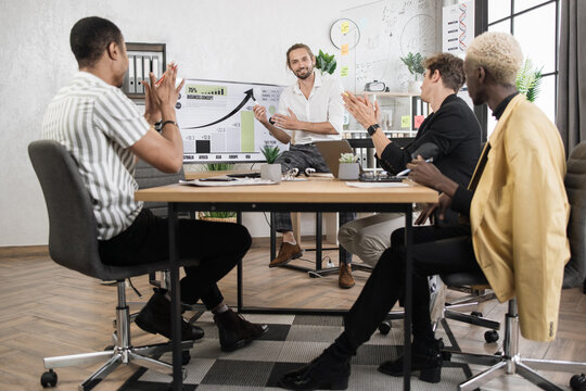 Young caucasian businessman in formal wear pointing on glass board with charts and graphs during working conference. Multi ethnic colleagues sitting at desk and listening male partner.