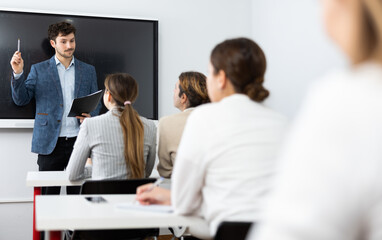 Young male teacher near interactive board, gesturing with hand while consecrating topic of lesson for adults in classroom