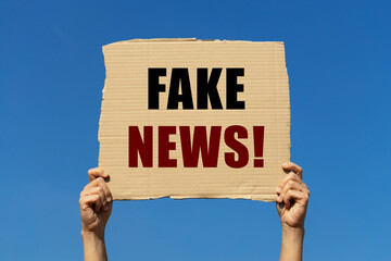 Fake news text on box paper held by 2 hands with isolated blue sky background. This message board...
