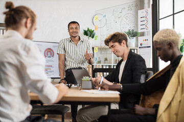 African man standing near big monitor and glass flip chart showing thumb up celebrating success while leading conference for multiracial colleagues.