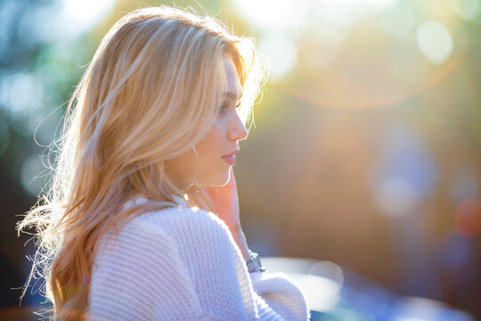 Close Portrait Of A Beautiful Young Caucasian Blonde Girl Dreamily Looking Right Wearing White Sweater With Blurred Blueish Autumn City Background, Contrasty Split Sun Lighting With Flare