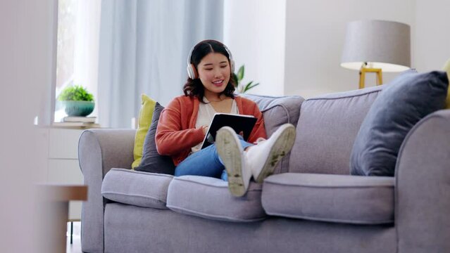 Tablet, headphones and woman relax on couch with music streaming app, social media and watching videos at home. Gen z asian person or young Korea girl on sofa, digital tech and listening to audio