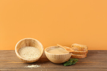 Bowls with tasty tahini, sesame seeds, bread and spinach on wooden table near color wall