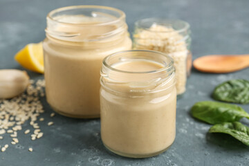 Obraz na płótnie Canvas Glass jars with tasty tahini, sesame seeds and spinach on dark background, closeup