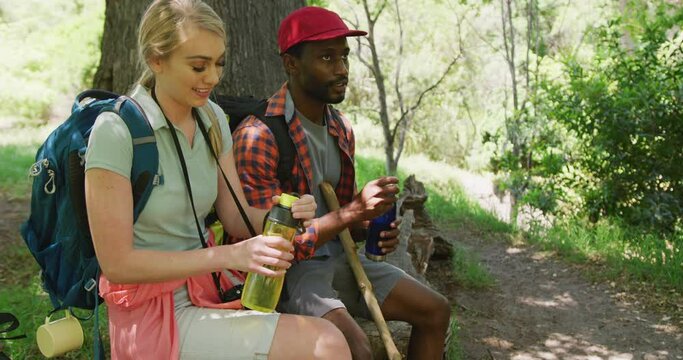 Happy diverse couple with backpacks sitting and drinking water in park, slow motion