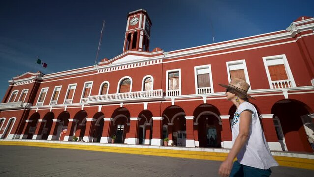 Happy smiling mature senior woman walking in front of the Municipal Palace in Motul, Yucatan, Mexico wearing ethnic attire, straw hat and sunglasses. Adventure is ageless.