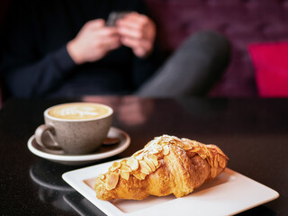 Croissant with flaked almonds on a white plate and a cup of coffee on the table in the cafe and a person holding smartphone on the background, selective focus