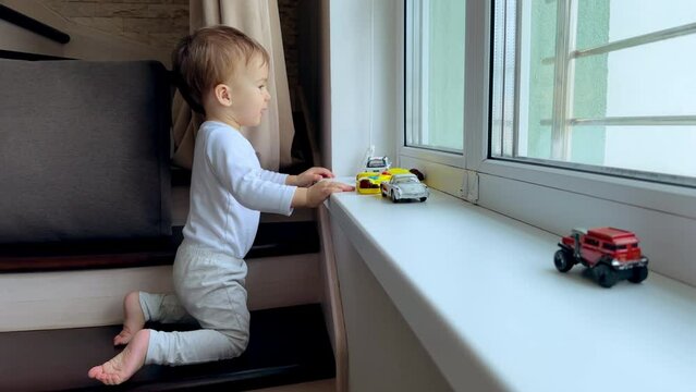 One year old toddler boy playing at the window. Little kid touches the toy cars placed on the window-sill.