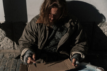 a homeless man sits on the street and writes on a sign