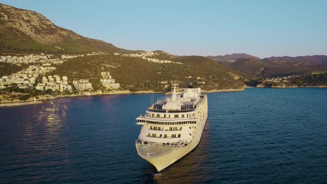 Huge white liner anchored in wide sea bay near a seaside town against backdrop of blue sky and mountainous coastline overgrown with green vegetation. View of the bow of the liner from a flying drone