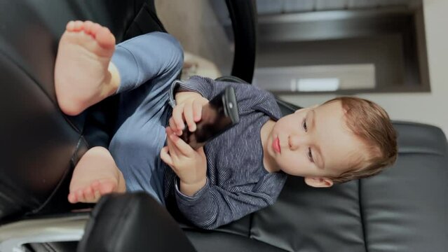 Peaceful Caucasian toddler sitting in the black office chair. Baby boy holds a tv remote and moves his tiny feet. Vertical screen.