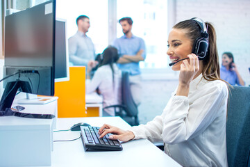 Smiling hotline operator woman with headset using computer in office. - obrazy, fototapety, plakaty