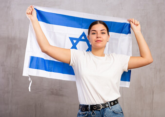 Portrait of serious patriotic young female holding national flag of Israel against gray wall...