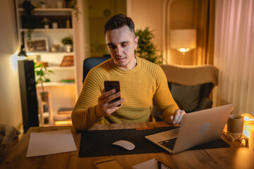 A young man is working late or studying at his home office while drinking coffee and using his...