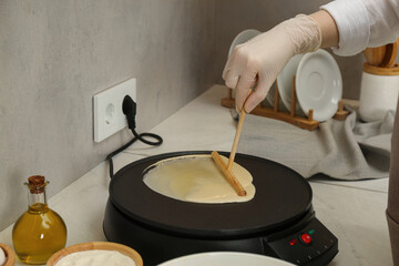Woman cooking delicious crepe on electric pancake maker at white marble table in kitchen, closeup