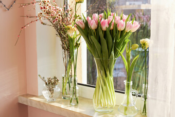 Many different spring flowers and branches with leaves on windowsill indoors